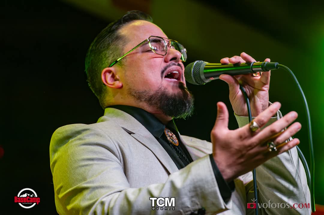 Lead vocalist in a cream blazer singing a ballad with his hand outstretched